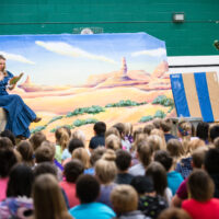 Performers from Lyric Opera of the North’s "Little LOON" school tour during the 2018-2019 season. In a school gym, a soprano in a blue pioneer dress sings while holding a book, seated before a desert backdrop. On the right, a baritone in cowboy attire and a black hat peeks playfully from behind a large prop cactus. A large audience of elementary students is shown from behind, watching the performance.