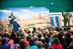 Performers from Lyric Opera of the North’s "Little LOON" school tour during the 2018-2019 season. In a school gym, a soprano in a blue pioneer dress sings while holding a book, seated before a desert backdrop. On the right, a baritone in cowboy attire and a black hat peeks playfully from behind a large prop cactus. A large audience of elementary students is shown from behind, watching the performance.