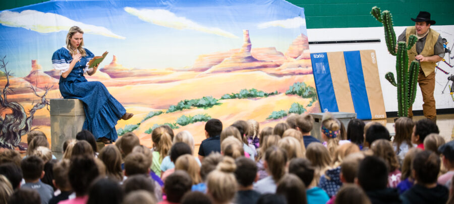Performers from Lyric Opera of the North’s "Little LOON" school tour during the 2018-2019 season. In a school gym, a soprano in a blue pioneer dress sings while holding a book, seated before a desert backdrop. On the right, a baritone in cowboy attire and a black hat peeks playfully from behind a large prop cactus. A large audience of elementary students is shown from behind, watching the performance.